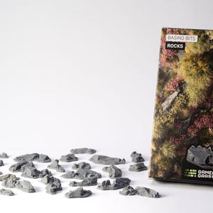 May include: A selection of small, grey model rocks scattered on a white surface, alongside a box with a detailed landscape design. The box is labelled "Basing Bits Rocks" and "Gamers Grass". The rocks are likely for miniature gaming terrain.