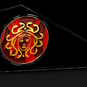 Peut inclure: Une plaque noire angulaire présentant un insert circulaire avec un relief doré représentant un visage aux cheveux en forme de serpents. Le fond de l'insert circulaire est divisé en sections verticales de rouge foncé et de rouge vif.