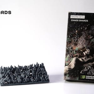 Puede incluir: Una base rectangular negra con muchas formas pequeñas, puntiagudas y de color gris oscuro, posiblemente terreno en miniatura. El producto está etiquetado como "Basing Bits Chaos Shards" y está junto a una caja con un diseño similar y el logotipo de "Gamers Grass".