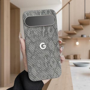 Puede incluir: Una funda de teléfono gris con un patrón de piel de serpiente. La funda tiene un recorte rectangular para la cámara en la parte superior y un logotipo "G" blanco en el centro. La funda está siendo sostenida.