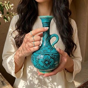 Pode incluir: Um jarro de cerâmica turquesa com gargalo estreito, alça curva e corpo arredondado decorado com motivos florais pretos pintados à mão. O recipiente tem um acabamento brilhante e uma borda alargada com linhas escuras detalhadas.