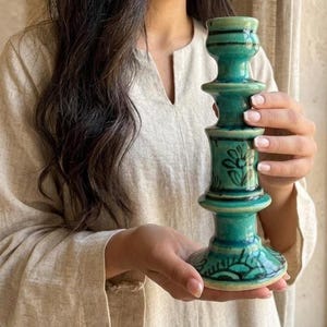 Può includere: Un portacandele in ceramica color verde acqua con finitura lucida e silhouette a livelli sovrapposti. La superficie presenta motivi floreali e a foglia neri dipinti a mano sulla base e sulla parte centrale, con un'ampia base circolare.
