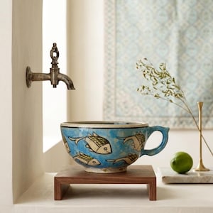 Op de afbeelding: Een keramische kom met een handvat, voorzien van een helderblauw glazuur versierd met handgeschilderde zwart-witte vismotieven. De kom staat op een rechthoekige houten standaard, direct onder een aan de muur gemonteerde metalen waterkraan.