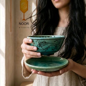 Op de afbeelding: Een turquoise keramieken kom en bijpassende schotel met een handgeschilderde zwarte visillustratie. De kom heeft een craquelé glazuurafwerking en een donkere rand. Het logo van NOOR STUDIO is zichtbaar met een icoon van een vis.