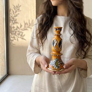 Puede incluir: Un candelabro de cerámica amarilla con motivos de peces pintados a mano en azul y negro en el cuerpo y un patrón floral alrededor de la base. La pieza tiene una base ensanchada, un cuello estrecho y una pequeña abertura circular superior.