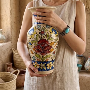 Könnte beinhalten: Eine Keramikvase mit cremefarbener Basis und handgemaltem Blumenmuster in Dunkelrot, Gelb, Grün und Blau. Die Vase hat einen weit ausgestellten Rand und einen verjüngten Hals, verziert mit horizontalen Bändern aus gelben und blauen Blüten.