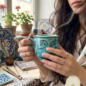 Puede incluir: Una taza de cerámica turquesa con un acabado de esmalte craquelado e ilustraciones de peces dibujadas a mano en negro. La taza presenta un asa resistente y una forma redondeada. Un pequeño logotipo muestra el texto NOOR STUDIO con un icono de ánfora.