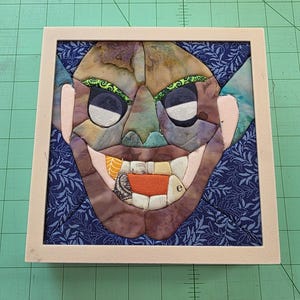 May include: A framed fabric art piece featuring a goblin face. The goblin has green eyes with black pupils, a wide mouth with teeth, and a textured, multi-colored face. The background is blue with a floral pattern, and the frame is a light beige color.