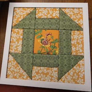 May include: A square fabric quilt block framed in white, featuring a central orange panel with a small illustration of a figure in a basket. The design includes alternating blocks of yellow floral fabric and green geometric patterned fabric.