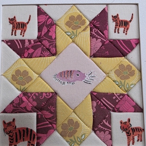 May include: A square fabric quilt block featuring a central diamond with a pink fish illustration, surrounded by yellow floral fabric, deep magenta patterned fabric, and four corner squares each displaying a small orange tiger illustration.