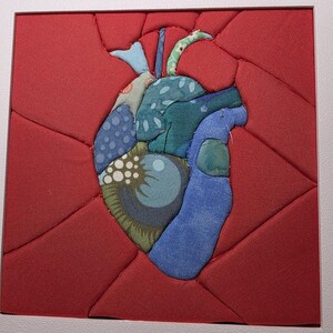 Puede incluir: Obra de arte textil enmarcada que representa un corazón humano estilizado. El corazón está hecho de retazos de tela en tonos azules, verdes y estampados, sobre un fondo rojo acolchado. La obra está encerrada en un marco blanco.