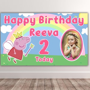May include: A rectangular birthday banner featuring a light blue sky with a rainbow, white clouds, and a yellow sun. It displays the text Happy Birthday Reeva 2 Today, a cartoon pig character with a crown and wand, and an oval photo of a young child.