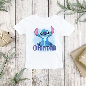 May include: A white short-sleeved t-shirt featuring a graphic print of the blue alien character Stitch sitting down. Below the character, the word OHANA is printed in a bold, purple, sans-serif font against a light blue watercolor background.