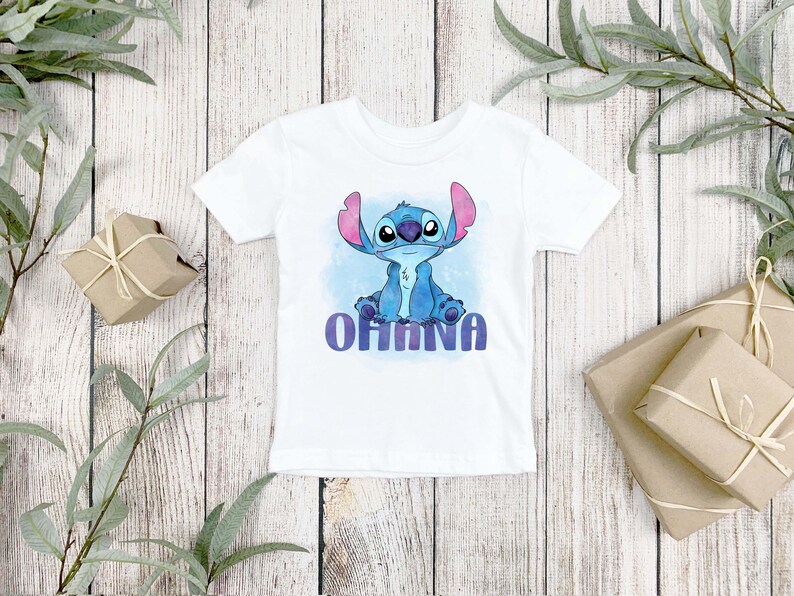 May include: A white short-sleeved t-shirt featuring a graphic print of the blue alien character Stitch sitting down. Below the character, the word OHANA is printed in a bold, purple, sans-serif font against a light blue watercolor background.