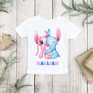 May include: A white short-sleeve t-shirt featuring a watercolor-style graphic of two cartoon characters, one pink and one blue, embracing each other. Below the characters, the name HALLIAH is printed in bold, alternating pink and blue capital letters.