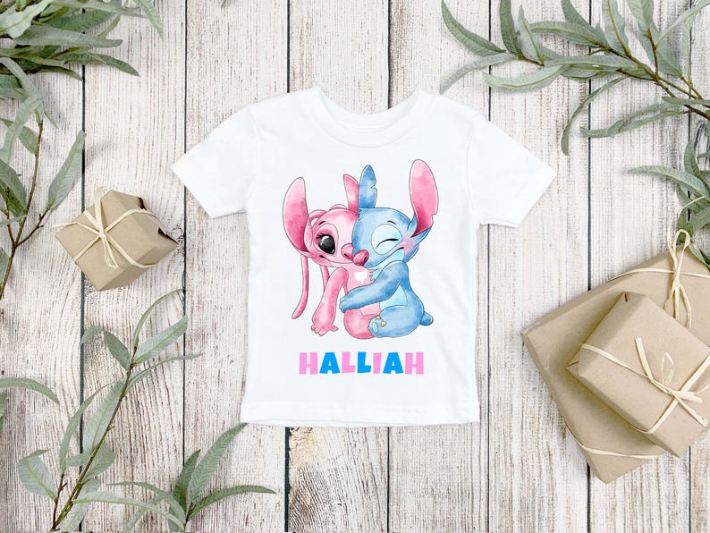 May include: A white short-sleeve t-shirt featuring a watercolor-style graphic of two cartoon characters, one pink and one blue, embracing each other. Below the characters, the name HALLIAH is printed in bold, alternating pink and blue capital letters.