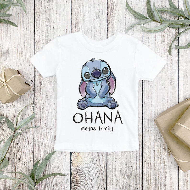 K&ouml;nnte beinhalten: Ein wei&szlig;es kurz&auml;rmeliges T-Shirt mit einer Aquarell-Illustration der blauen Figur Stitch im Sitzen. Unter der Figur steht der Text OHANA in gro&szlig;en schwarzen Buchstaben, gefolgt von dem kleineren schwarzen Text means family.