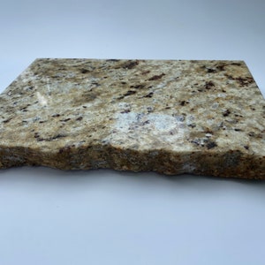 May include: A rectangular slab of light brown granite with a rough, uneven edge. The granite has a speckled pattern of darker brown and black flecks.