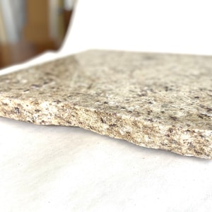 May include: A close-up of a light brown granite tile with a rough, uneven edge. The tile has a speckled appearance with small, dark brown flecks throughout.