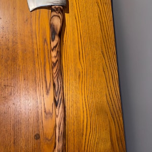 May include: A single-bit axe featuring a polished silver metal head and a long, curved wooden handle. The handle displays a distinct, dark, flame-scorched wood grain pattern that runs vertically along the entire length of the grip.