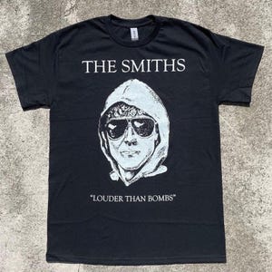 Op de afbeelding: Zwart t-shirt met de bandnaam "THE SMITHS" in het wit boven een witte afbeelding van een persoon met een zonnebril en een capuchon. De zin "LOUDER THAN BOMBS" staat eronder afgedrukt.