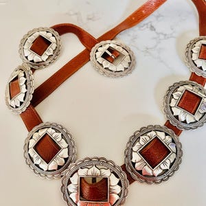 May include: A brown leather belt with silver-toned decorative medallions. Each medallion has a scalloped edge and a brown leather center. The belt buckle is also silver-toned with a matching leather insert. A stylish accessory.