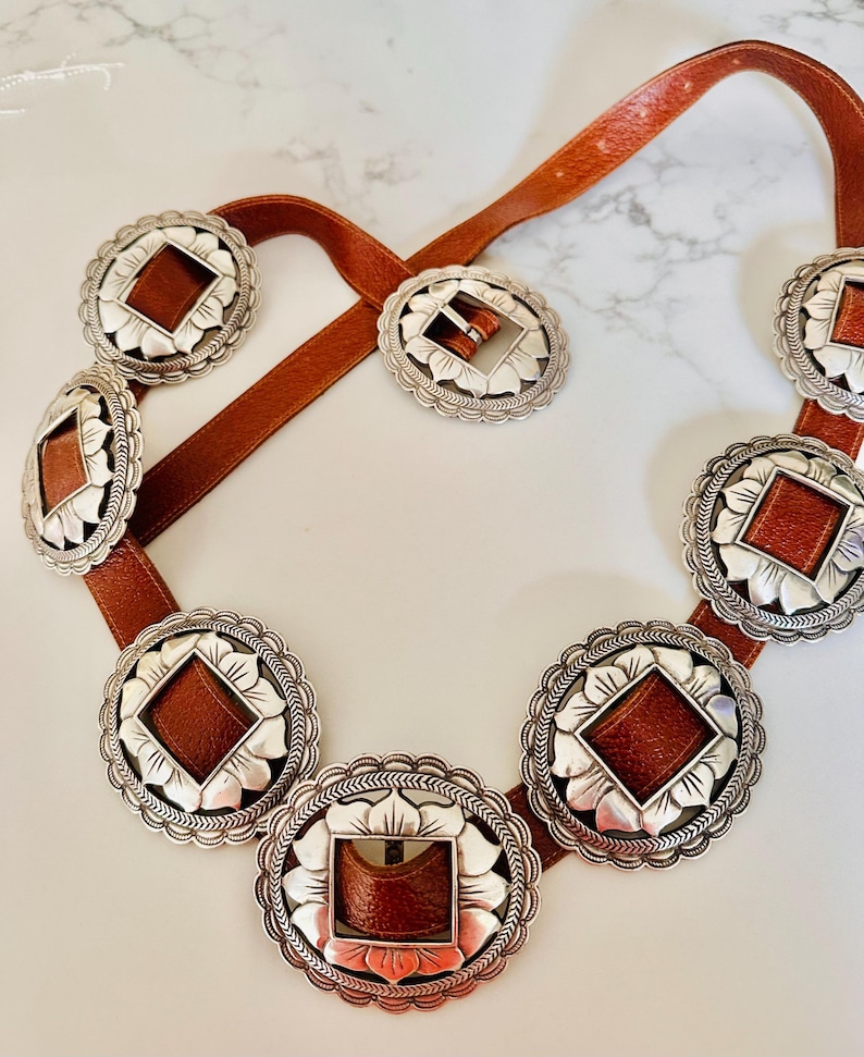 May include: A brown leather belt with silver-toned decorative medallions. Each medallion has a scalloped edge and a brown leather center. The belt buckle is also silver-toned with a matching leather insert. A stylish accessory.