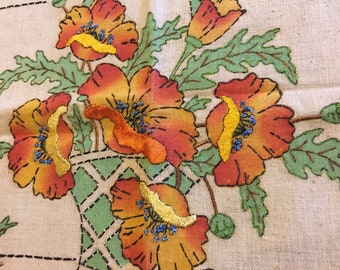 May include: Embroidered floral design featuring orange and yellow poppies with green leaves and stems. The flowers are arranged in a green basket-like shape, with blue accents in the center of the flowers. The embroidery is on a cream-colored fabric.
