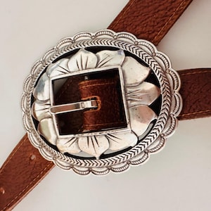 May include: A brown leather belt with a large, ornate silver buckle. The buckle has a floral design around the edge and a square center. The belt is a medium brown color and appears to be made of leather.