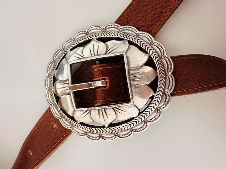 May include: A brown leather belt with a large, ornate silver buckle. The buckle has a floral design around the edge and a square center. The belt is a medium brown color and appears to be made of leather.