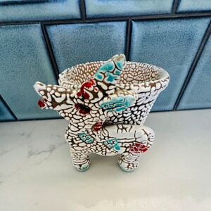 May include: A ceramic planter shaped like a four-legged animal with a textured white surface featuring deep brown cracks. The design includes accents of bright turquoise and red glaze on the ears, body, and legs, creating a distinct contrast.