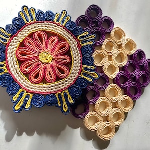 May include: A circular woven mat featuring a central coral and yellow flower motif surrounded by blue and yellow coiled patterns, partially overlapping a square grid of interconnected purple and beige woven rings made of coiled paper strips.