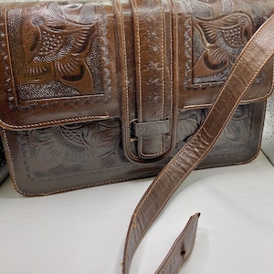 May include: A brown leather satchel with embossed floral and bird designs. The bag features a long shoulder strap and a buckle closure. The leather has a distressed appearance, with visible stitching and wear.