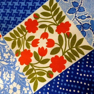 May include: Close-up of a patchwork fabric with various patterns. The design includes squares with floral, polka dot, and geometric patterns in shades of red, blue, green, and white. The fabric has a vintage aesthetic.