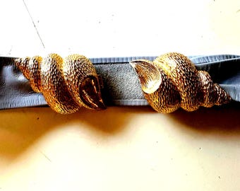 May include: A belt featuring two large, textured gold-tone metal seashell-shaped buckles that meet in the center. The belt strap is made of a smooth, light gray material that extends behind the metallic shell ornaments on both sides.