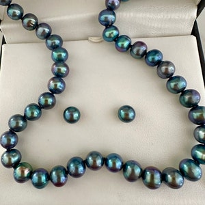 May include: A dark gray pearl necklace and matching stud earrings set. The pearls have a lustrous sheen with hints of green and purple. The jewelry is displayed in a white box with a black interior.