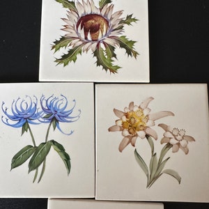 May include: Four ceramic tiles with painted floral designs. The top tile features a white flower with green leaves and a brown center. The left tile features two blue flowers with green leaves. The right tile features a white flower with yellow centers and green leaves. The bottom tile features three purple flowers with green leaves.