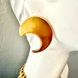 May include: A pair of large, gold-tone crescent moon shaped earrings. The earrings are made of a smooth, shiny metal.