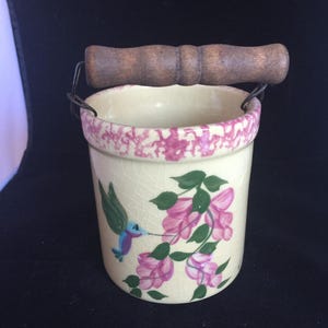 May include: A cylindrical ceramic crock with a cream-colored crackle glaze, featuring a hand-painted hummingbird and pink flowers. The rim has a pink speckled pattern, and the vessel includes a dark brown wooden handle attached by wire loops.