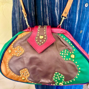 May include: A colorful leather handbag with a shoulder strap. The bag features a patchwork design in brown, green, pink, and tan, embellished with gold-colored studs. The bag has a burgundy flap with gold studs.