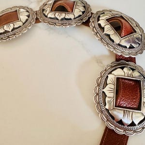 May include: A brown leather belt with large, ornate silver-toned buckles. Each buckle has a square, brown leather insert and a decorative floral design around the edges. The belt is displayed on a white surface.