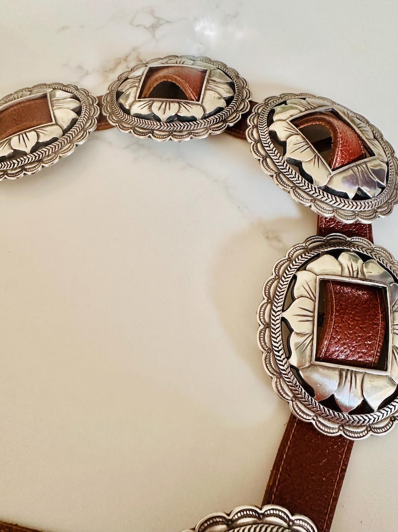 May include: A brown leather belt with large, ornate silver-toned buckles. Each buckle has a square, brown leather insert and a decorative floral design around the edges. The belt is displayed on a white surface.