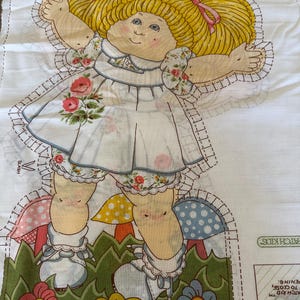 May include: A vintage Cabbage Patch Kids fabric panel featuring a doll illustration. The doll has blonde pigtails with pink bows, a white dress with floral accents, and is standing in a garden scene with mushrooms and flowers.