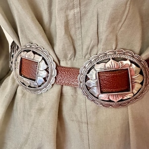 May include: A close-up of a brown leather belt with two large, silver-colored oval buckles. Each buckle has a rectangular brown leather center and a decorative scalloped edge. The belt is worn over a light green garment.