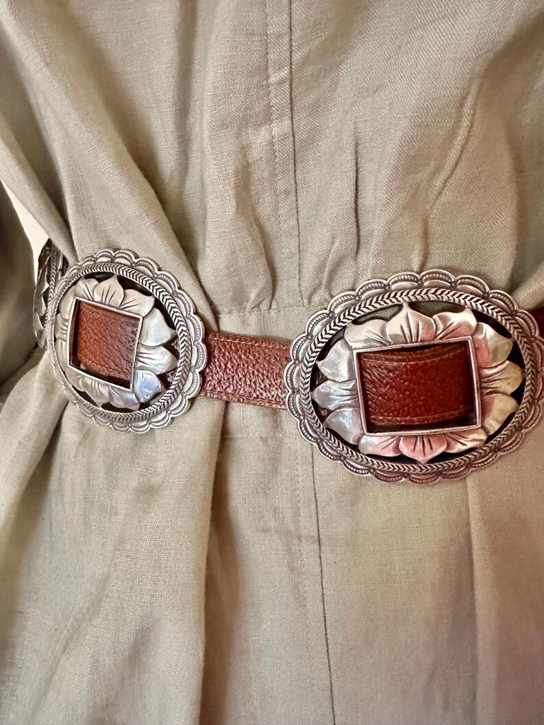 May include: A close-up of a brown leather belt with two large, silver-colored oval buckles. Each buckle has a rectangular brown leather center and a decorative scalloped edge. The belt is worn over a light green garment.