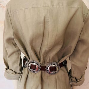 May include: A brown leather belt with two large, ornate silver buckles. The buckles have a scalloped edge and a square, brown center. The belt is worn over a light green linen jumpsuit.