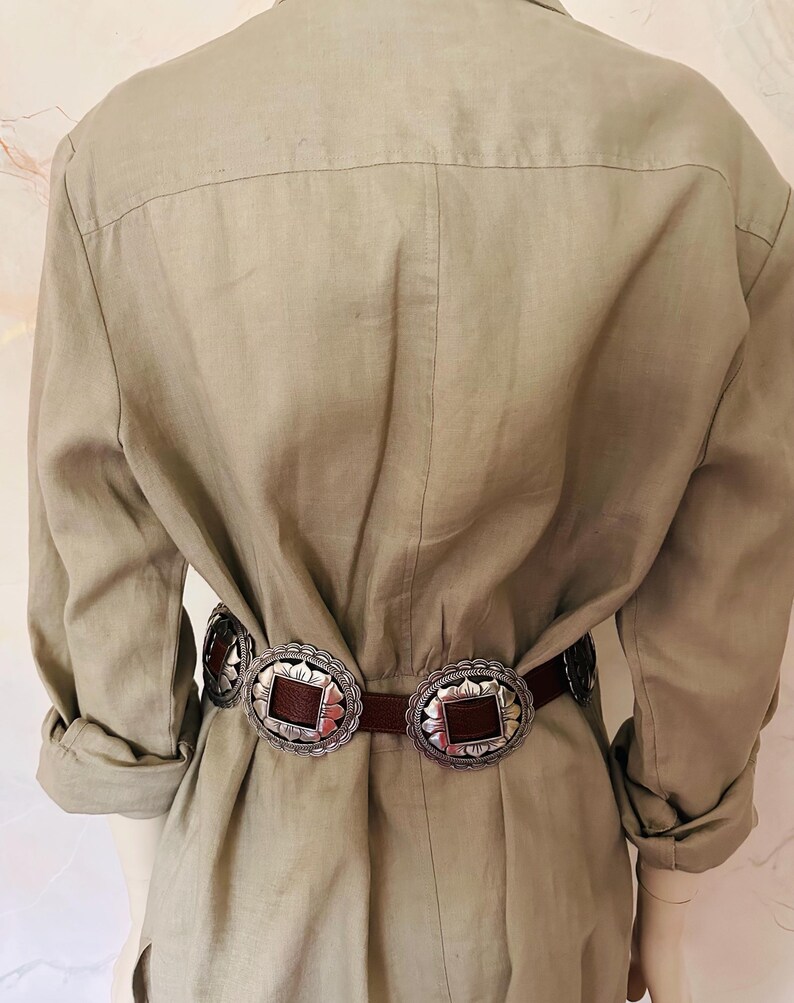 May include: A brown leather belt with two large, ornate silver buckles. The buckles have a scalloped edge and a square, brown center. The belt is worn over a light green linen jumpsuit.