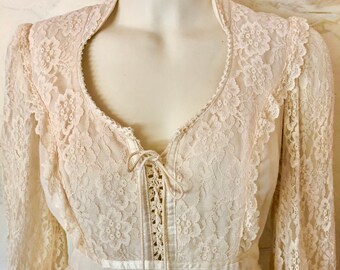 May include: Cream-colored vintage dress featuring a delicate lace overlay. The dress has long sleeves, a high neckline, and a bodice with a lace-up detail. A satin ribbon accents the waist, and the skirt is a flowing, light fabric.