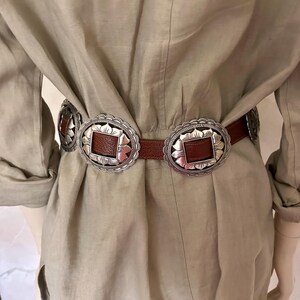 May include: A brown leather belt with silver-toned decorative buckles. The buckles have a square center and a scalloped edge. The belt is worn over a light tan dress.