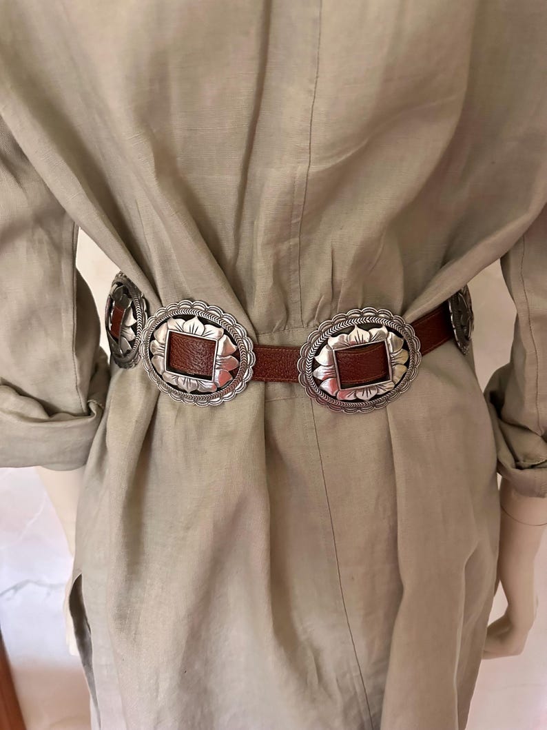 May include: A brown leather belt with silver-toned decorative buckles. The buckles have a square center and a scalloped edge. The belt is worn over a light tan dress.
